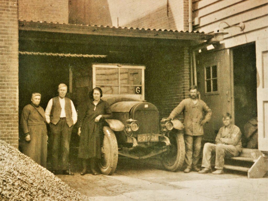 De familie Nat met vrachtauto naast pakhuis De Hoop (circa 1927). Van links naar rechts Trijntje Nat – Beers, haar echtgenoot Dirk Nat, dochter Gezina en zoons Jan en Cor. Links ligt een hoop kolen bestemd voor de verwarmingsinstallatie en de zuiggasmotor van de fabriek. De vrachtauto was een Fiat of een Martini, die met aanslingeren gestart moest worden.