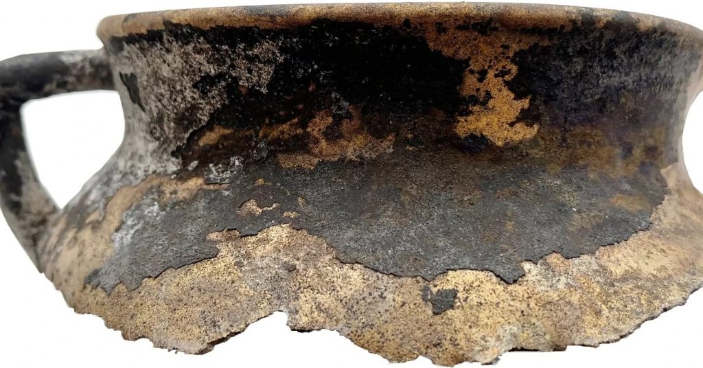 De rand van een bronzen kookpot met nog één oor dat eraan vast is gesoldeerd. Het is een bolvormige kookpot geweest, op drie pootjes en met twee oren onder de rand, een zogeheten grape.