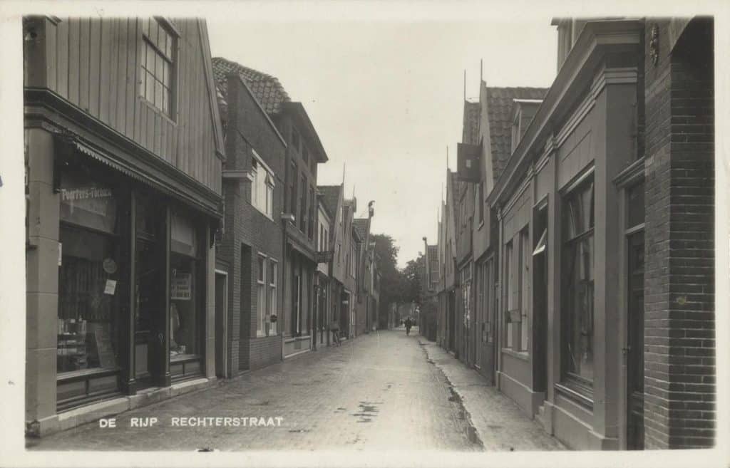 Prentbriefkaart De Rijp Rechtestraat. Circa 1933.  In de verte steekt het Huis met de Arend tegen de lucht af. In de tabakszaak hangt in de etalage een bord; de tekst is niet helemaal zichtbaar maar er staat waarschijnlijk ‘Koopt Elders Niet Wat Eigen Dorp U Biedt’. Men zat toen midden in de crisisjaren. Foto D. van der List. Collectie Kaptein