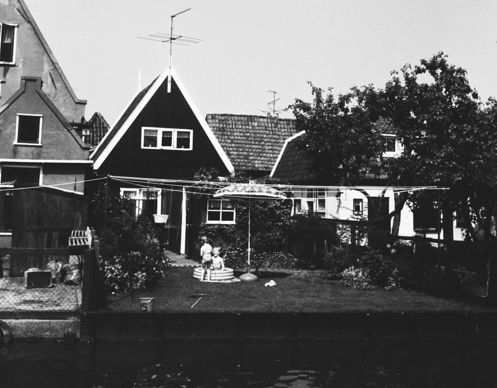 Zicht op de achtertuin van Oosteinde 16 in De Rijp. Gezien vanaf het water. In de tuin twee kinderen op een zomerse dag in een badje. Jaren 1970.