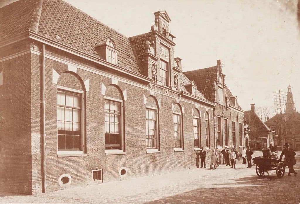 Het Huis van Achten in 1903. Zuidelijke gevel van het in 1656 gestichte Huis van Achten of Provenhuis van Nordingen. De ingang van het hofje is in de Lombardsteeg 23. Links achter de personen de gevel van het Hof van Sonoy. Voor de Waagtoren op de achtergrond een telefoonmast van Pot.