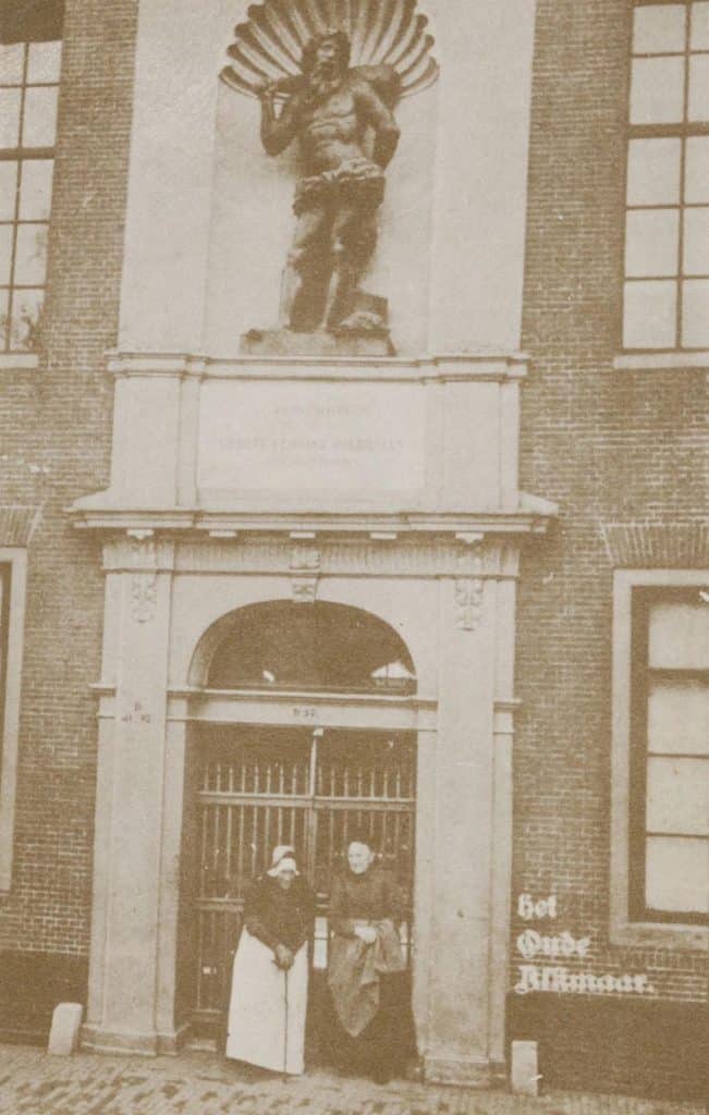 Prentbriefkaart. Het Wildemanshofje De poort met vermoedelijk twee bewoonsters in klederdracht in 1905.