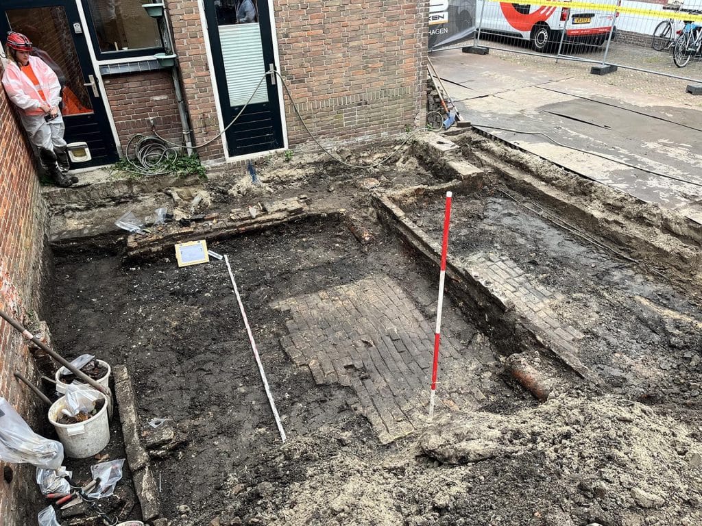 Opgraving aan Verdronkenoord 66 naast de Laurentiuskerk. De bakstenen werkplaatsvloer die werd blootgelegd tijdens de opgraving. De ronde schijf pek of teer is tijdens het verwijderen van de kleilaag meegekomen en ligt in het stort voor de rood/witte jalonstok. Kijkrichting: ZZW (richting het Verdronkenoord).