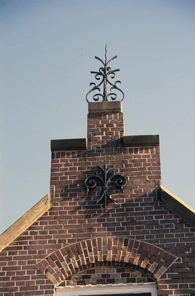 Detail van de nok Brugwachterswoning met tuitgevel aan de Eilandswal. De trapgevel aan de Eilandswal is extra rijk uitgewerkt. Met een band van gele baksteen in ruitvorm en een opvallende ijzeren bekroning bovenin. Boven de ramen zitten sierlijke segmentbogen met gekleurde accenten en subtiele details. Foto J. Elsinga 2003.