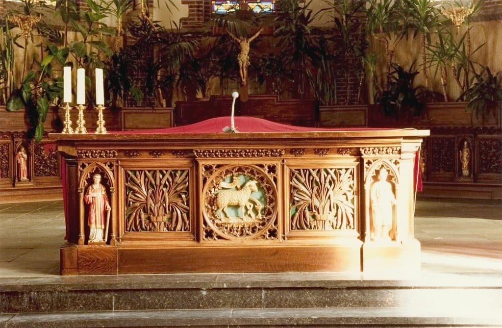 De rijkgesneden eikenhouten neogotische communiebanken (1881) uit het atelier van Margry werden in de jaren ‘70 vermaakt tot een nieuw hoofdaltaar. En tot de driedelige achterwand in het koor. Op de panelen zie je druiventrossen of korenaren. Afgewisseld met heiligenbeelden en voorstellingen die betrekking hebben op de Eucharistie. Foto J. Elsinga 1992.