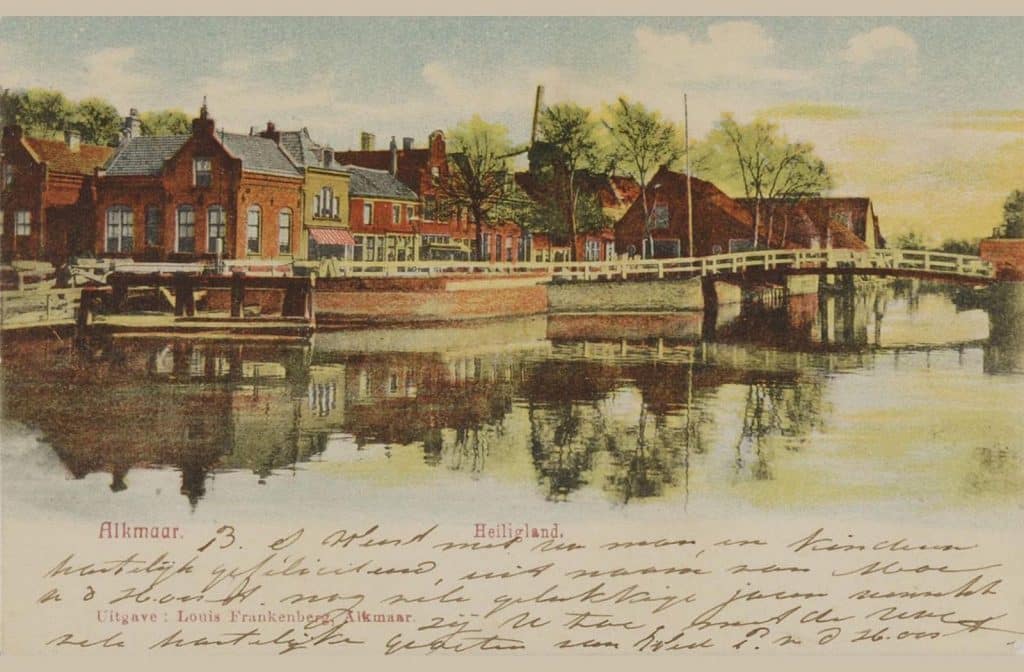 Prentbriefkaart van de Eilandsbrug voorheen de Jaagbrug. Links de Brugwachterswoning aan de Eilandswal. Gevelwand op het Schermereiland ofwel het Rooie Dorp. Links een stukje van de Schermer vlotbrug (1821-1958). Op de achtergrond molen de Wachter (1896-1918). Collectie prentbriefkaarten. Circa 1903.