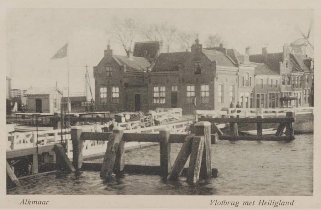 Prentbriefkaart van de Schermervlotbrug, een drijvende brug over het Noordhollands Kanaal. De vlotbrug lag in het verlengde van het Heiligland op het Schermereiland. Dit wordt ook wel het Rooie Dorp genoemd. De brug verbond de Eilandswal met de Achterweg ofwel de Wageweg met het centrum. Molen de Wachter (1896-1918) reikt boven de huizen uit.
De vlotbruggen, ontworpen door Jan Blanken, zijn gebruikt bij de aanleg van het Noordhollands Kanaal in 1819-1824. De Schermervlotbrug lag er van 1924-1958.
De Achterweg heette vanaf 12-4-1973 de Wageweg. Collectie prentbriefkaarten. Circa 1910.