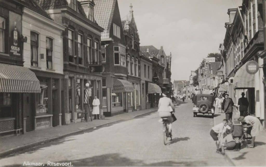 Prentbriefkaart van het straatbeeld in het Ritsevoort kapper H Zinger staat voor zijn zaak Circa 1937