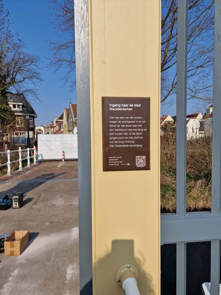 Dit bordje met informatie over het hek en een QR code. Door de code te scannen kom je op de website van Erfgoed Alkmaar. Daar vind je een pagina met informatie, verhalen, foto's en filmpjes over het hek.
