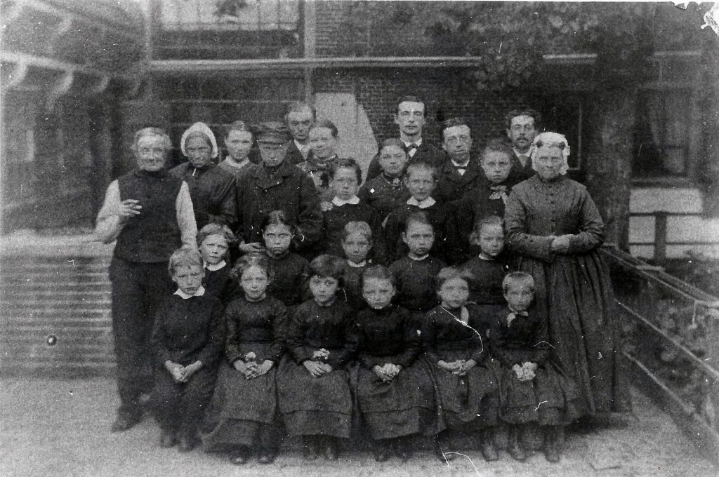 Een oude zwart/wit foto van een groep Weeskinderen van het Algemene Wees- en Armenhuis. Met links de weesvader, rechts de weesmoeder en achter de kinderen de regenten.