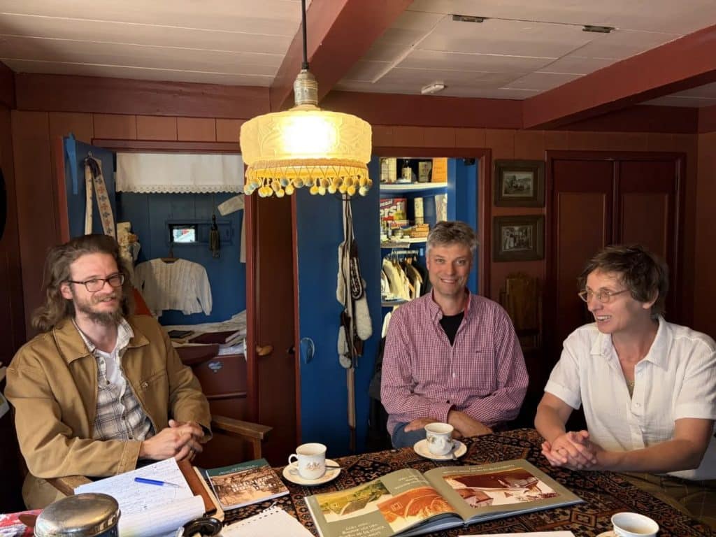 In de woonkamer van het huisje zitten drie mensen aan de eettafel. Er staan kopjes en er liggen boeken op tafel. In de donkerrode achterwand zie je een bedstee.