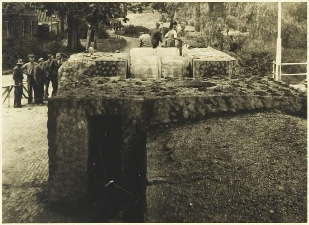 Op de voorgrond de spermuur. Links naast de muur staan 5 mannen. Achter de muur staan 6 mannen. Helemaal links zie je de zijmuur van het Hofje van Paling en Van Foreest. Bovenaan in het midden het wandelpad over het Bolwerk.