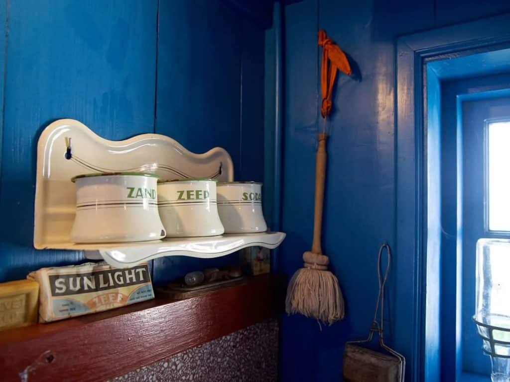 Een plankje bevestigd aan een blauwe houten wand. Met 3 potten, voor zand, zeep en soda. Daaronder een oude verpakking van SUNLIGHT zeep.