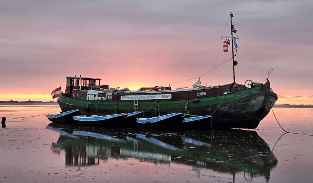 De lucht is roze, geel en lila. Het groen/zwarte schip ligt aangemeerd. Er hangen 4 kleine blauwe zeilboten aan het schip vast.