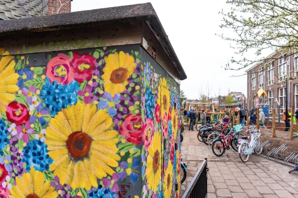 Op de voorgrond een met bloemen beschilderde fietsenschuur. Op de achtergrond spelende kinderen op de nieuwe speeltoestellen.
