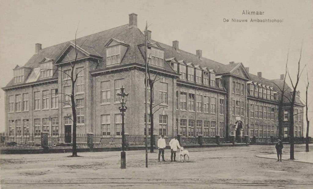 Ambachtsschool en Burger Avondschool. Gezien vanaf de hoek van de Stationsweg en het Scharloo. In het midden staan 2 mannen en een hond. Rechts staat een persoon met een pet op.