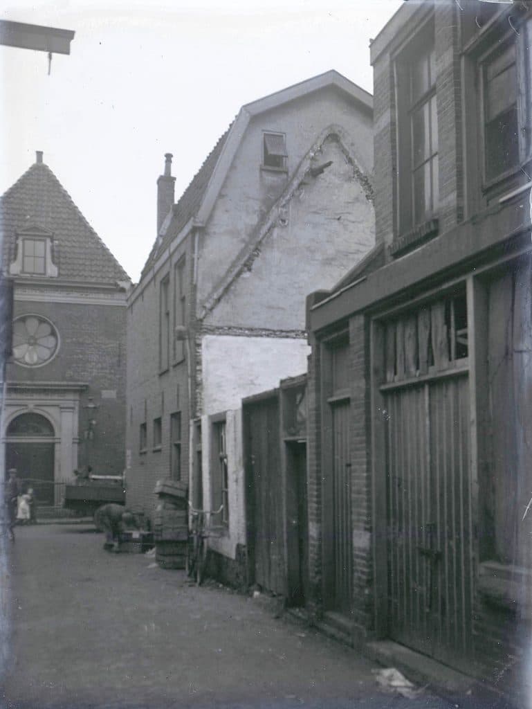 Foto uit 1926 van de Synagoge. Links zie je een vader met 2 kinderen. Voor een van de huizen staat een man die bezig is met kisten. Er staat een fiets voor een wit huis.