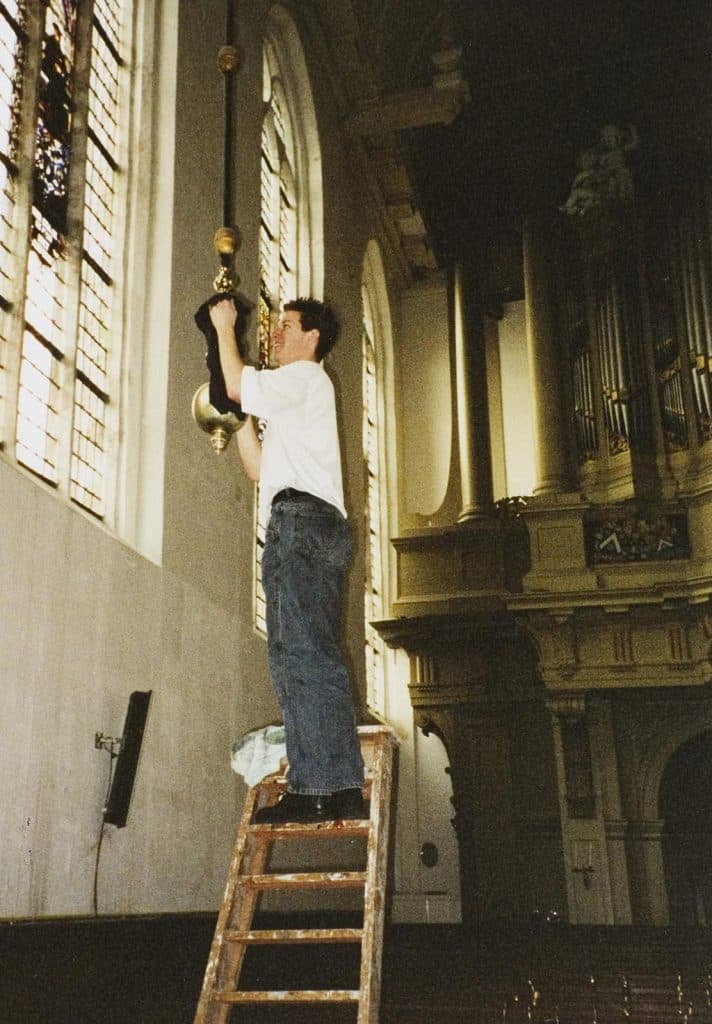 Een vrijwilliger staat op een houten ladder en poetst het koper van de kroonluchter.