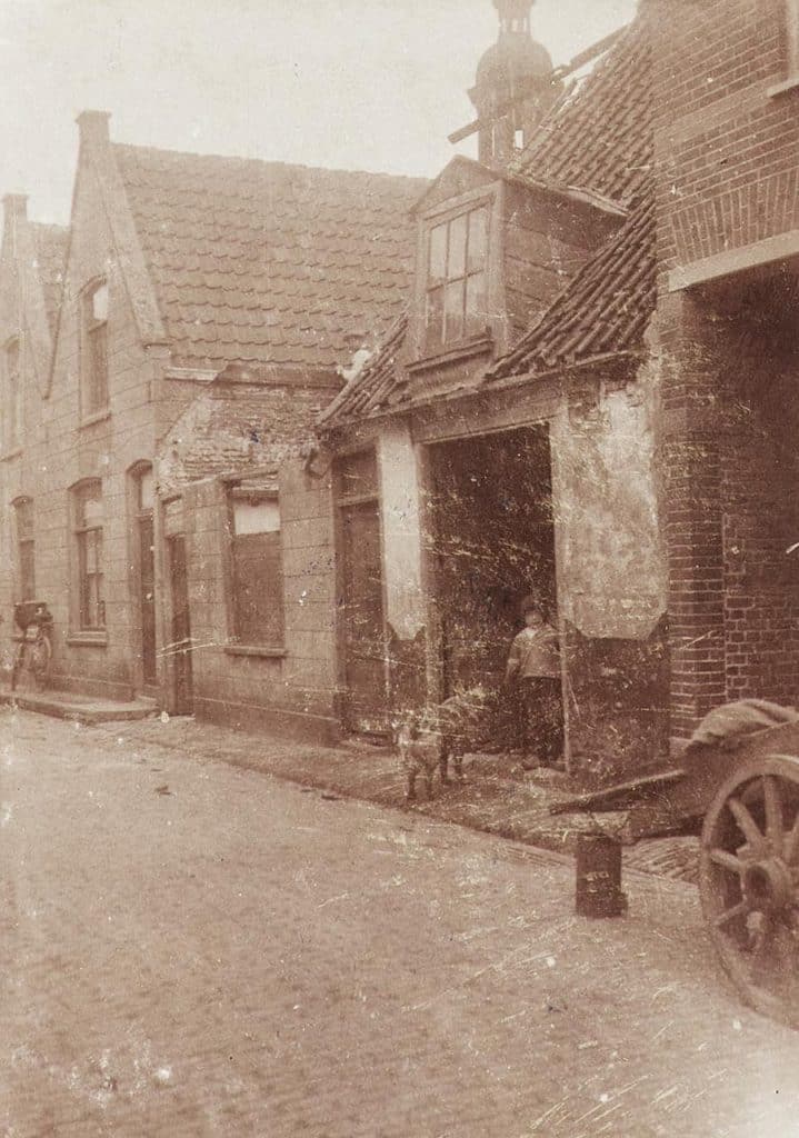Oude foto met op de voorgrond een handkar. In de deuropening van een van de huizen staat een kindje met een dier. Op het dak van het middelste huis staat eenjongetje met een wit hemd en een pet op.